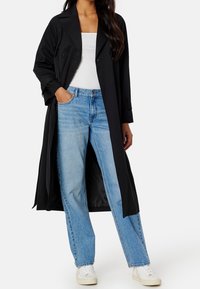 Černý dlouhý kabát s cíleným střihem, dvojřadé knoflíky, nošený přes bílý top a světle modré džíny. Hladká textura látky.