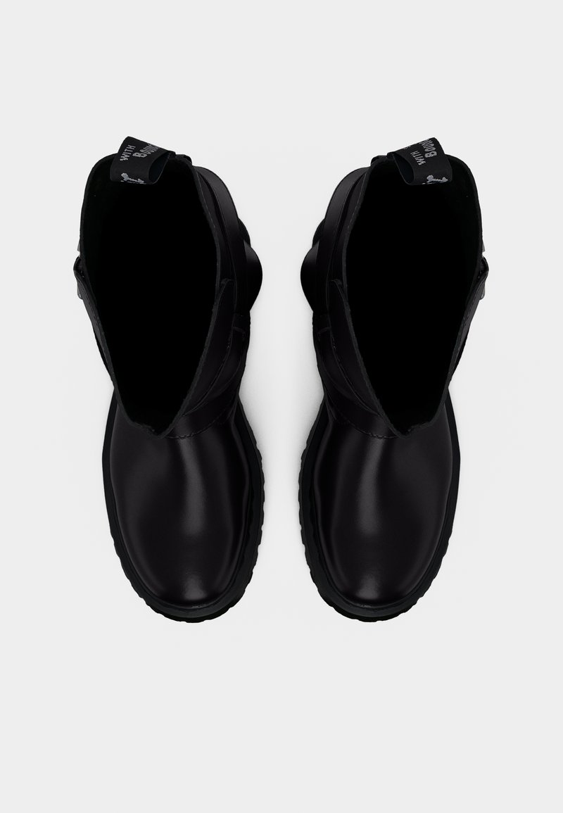 Coppia di stivaletti in pelle nera con punte arrotondate, suole spesse e linguette posteriori, visti dall'alto su sfondo bianco.