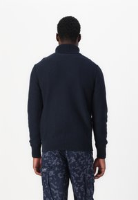 Marineblå strikkegenser med høy krage, ribbestrikkede mansjetter og kant. Brukt sammen med mønstrede cargobukser som viser et teksturert design.