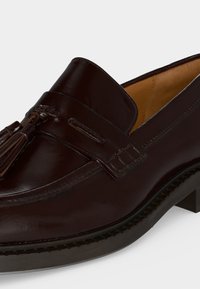 Σκούρος καφέ δερμάτινος loafer με λεπτομέρεια φουντίτσας, λείο φινίρισμα, ραμμένα άκρα και χαμηλό τακούνι ορατό σε μονόχρωμο φωτεινό φόντο.