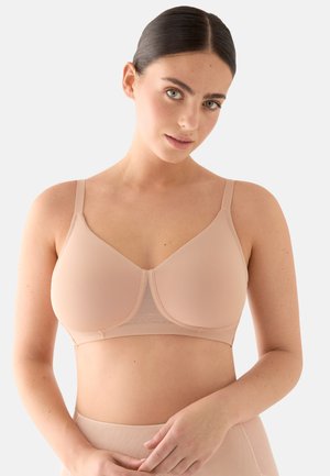 Mujer de cabello oscuro con un sujetador sin aros de color nude liso y ropa interior de cintura alta a juego, sobre un fondo claro.