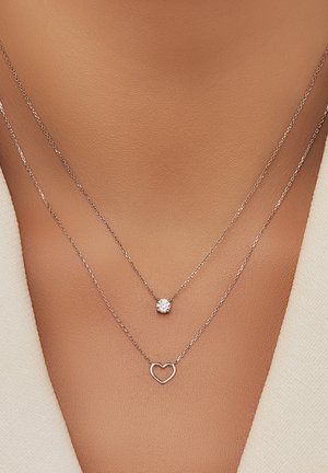 Colliers en argent superposés avec un pendentif en cristal rond et un charme en forme de cœur, reposant contre une surface de peau lisse.