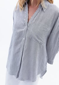 Camicia a maniche corte con bottoni a strisce blu e bianche, caratterizzata da una vestibilità ampia, taschino sul petto e un leggero orlo curvo.