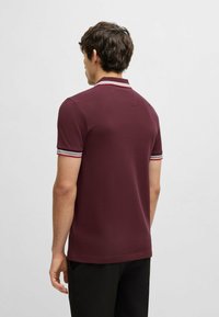 Burgundin värinen polo-paita, joka on valmistettu tekstuurista kankaasta. Paidassa on valko- ja punaraitainen kaulus, lyhyet hihat ja suora helma.