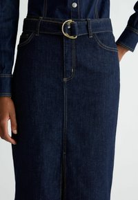 Gonna in denim blu scuro con un design aderente, dotata di una cintura larga con una fibbia circolare in argento e cuciture gialle a contrasto.