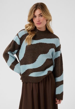 Femme aux longs cheveux blonds portant un pull marron à rayures abstraites bleu clair et une jupe marron foncé, debout devant un fond uni.