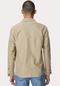 Beige katoenen jack met een kraag, lange mouwen en knoopsluiting bij de manchetten. Heeft een gladde textuur en een eenvoudig ontwerp, gezien vanuit de achterkant.