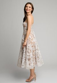 Sukienka bez ramiączek, sięgająca do kolan, w przezroczystym beżowym kolorze z białą koronką w kwiaty. Posiada dopasowaną górę i spódnicę w kształcie litery A, zestawiona z cielistymi szpilkami.