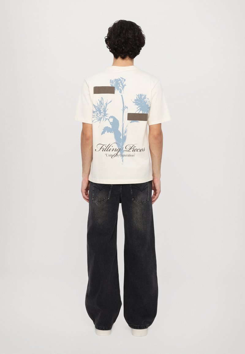T-shirt beige à manches courtes avec un design floral bleu et des accents marron au dos ; associé à un pantalon noir ample.