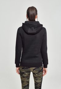 Femme aux cheveux bruns attachés en queue de cheval, vêtue d'un sweat à capuche noir et de leggings camouflage verts, debout dos à la caméra devant un fond uni.