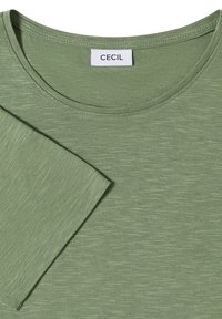 Chemise verte à manches courtes avec une texture subtile et un col rond, présentant une étiquette blanche avec le texte "CECIL" au niveau du col.