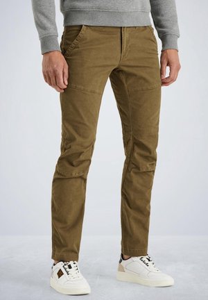 Mand iført brune slimfitbukser med knæsyninger, grå sweatshirt og hvide sneakers, stående på en grå gulv.