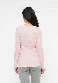 Top de manga larga color rosa claro con cinturón de satén envolvente en la cintura y textura acanalada, combinado con pantalones ajustados grises.