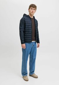 Polstret sort jakke med hætte, beige sweater, lyseblå jeans og beige sko. Har quiltet tekstur og ribbede manchetter.