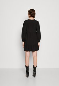 Vestito nero con design a strati, maniche lunghe e vita aderente. Presenta un tessuto morbido e un collo rotondo. Indossato con stivali neri alla caviglia.