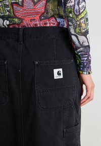 Sorte denim overaller med en lomme med Carhartt-logo, slitesterkt stoff og synlige sømdetaljer. Rikt tekstur og holdbart design.