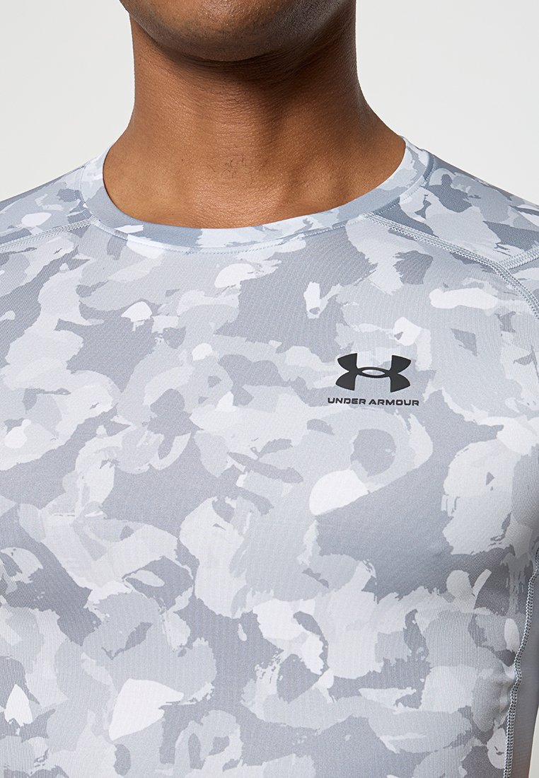 Camisa deportiva con patrón de camuflaje gris, de cuello redondo y manga corta. Presenta el logo de Under Armour en negro en el pecho. Textura suave.