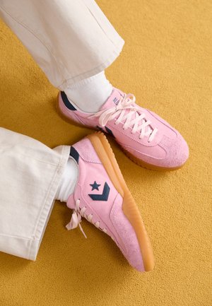 Pies con zapatillas de gamuza rosa con suelas de goma y logo de estrella negra, calcetines blancos y pantalones crema sobre una alfombra color mostaza.