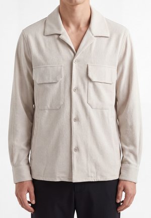 Man draagt een beige overhemd met knopen, lange mouwen, kraag en twee borstzakken, gecombineerd met een zwarte broek, staand tegen een witte achtergrond.