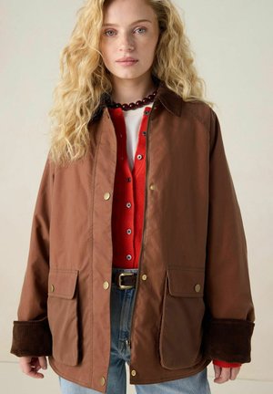 Brown jacket with a zip front and golden snap buttons. Features corduroy cuffs and two front pockets, layered over a red button-up.