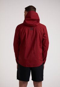 Rode waterdichte jas met een capuchon, met een gladde textuur en een logo op de achterkant. Gecombineerd met zwarte shorts.