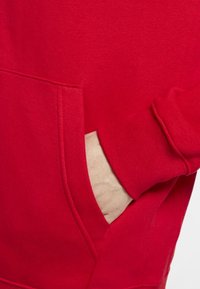 Röd sweatshirt med en framficka och ribbad mudd. Materialet verkar mjukt med en slät yta och sydda detaljer längs sömmarna.