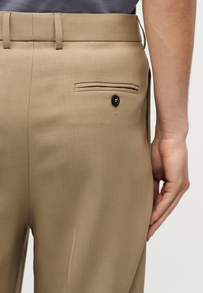 Gros plan sur une personne portant un pantalon beige sur mesure avec une poche arrière boutonnée, montrant une partie de la main reposant sur le côté.
