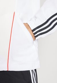 Chaqueta deportiva blanca con acentos en rojo y negro, que cuenta con un bolsillo con cremallera y puños acanalados con tres rayas negras.