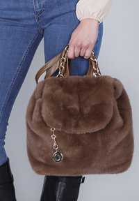 Donna che tiene in mano una borsa in finta pelliccia marrone con manici in pelle e catena, indossando jeans blu e stivali neri fino al ginocchio.