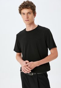 Sort kortærmet t-shirt med rund hals, lavet af et glat stof. Bæres med sorte bukser og et bælte, sat mod en lys baggrund.