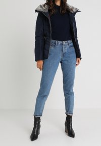 Giubbotto puffer blu navy con cappuccio foderato in pelliccia sintetica, abbinato a jeans a vita alta azzurri chiari e stivali neri alla caviglia. Design semplice e casual.