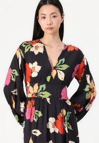 Blomstrete maxikjole i svart stoff med livlige røde, rosa og grønne mønstre. V-hals og lange ermer med samlet midje detaljer.