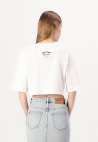 Balta, trumpa marškinėliai su plačiomis rankovėmis, ant nugaros turi juodą logotipo etikėtę. Sujungti su šviesiai mėlynomis, aukštu juosmeniu kelnių.