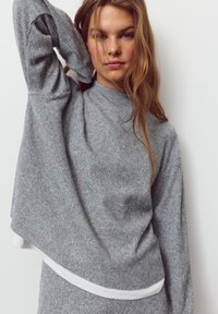 Pulover tricotat gri deschis cu guler înalt și mâneci lungi. Prezintă o tivă albă stratificată la bază și o croială lejeră.