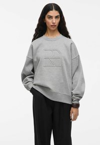Felpa grigia con un logo testurizzato sul davanti, maniche ampie e vestibilità rilassata. Abbinata a pantaloni neri e un accento marrone.