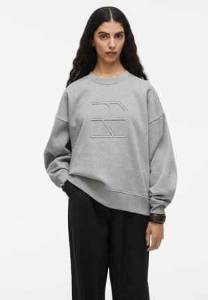 Grauer Sweatshirt mit einem strukturierten Logo auf der Vorderseite, weiten Ärmeln und einem lässigen Schnitt. Kombiniert mit schwarzen Hosen und einem braunen Akzent.