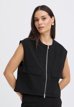 Donna con capelli lunghi e scuri che indossa un gilet nero senza maniche con cerniera frontale e due grandi tasche con patta, in piedi su uno sfondo chiaro.