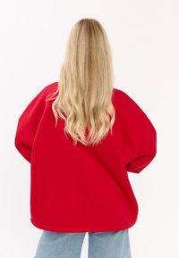 Czerwona, oversized bluza z opadającymi ramionami i ściągaczem u dołu, zestawiona z jasnoniebieskimi dżinsami. Miękka faktura, minimalistyczny design.