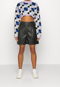 Pull en crochet court avec un motif à carreaux violet, noir, bleu et vert, associé à un short en cuir noir et des baskets blanches.
