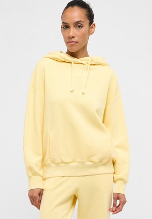 Woman wearing a pale yellow hooded sweatshirt and matching pants, standing against a plain light background.