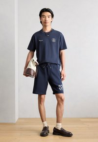 Άνδρας που φοράει μπλε ναυτικό T-shirt και σορτς με τον αριθμό "67", κρατώντας μια μπεζ και καφέ τσάντα, στέκεται σε ξύλινο δάπεδο μπροστά από έναν απλό φωτεινό τοίχο.