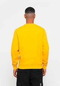 Yellow sweatshirt with long sleeves, crew neck, and ribbed cuffs. Visible stitching and a smooth texture. Worn with black cargo pants.