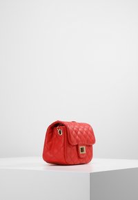 Borsa a tracolla rossa trapuntata con chiusura a patta, dettagli in metallo dorato, forma compatta e texture liscia. Progettata per trasportare gli essenziali.