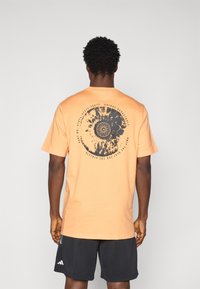 T-shirt en coton orange à manches courtes avec un design graphique circulaire noir et du texte au dos, avec un col rond et une coupe décontractée.
