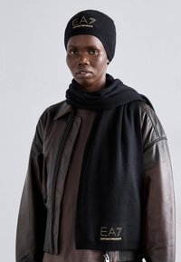 Svart strikket lue og skjerf-sett, med "EA7 Emporio Armani"-logo i gull. Båret med en brun t-skjorte og mørk lærjakke.