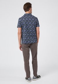 Camisa de manga corta en tela oscura con un patrón floral en azul y verde, combinada con pantalones texturizados en marrón claro y zapatillas grises.
