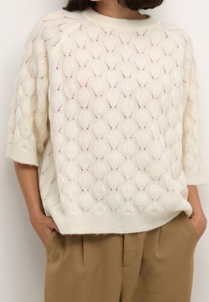 Femme portant un pull en tricot crème avec un motif de feuilles et des manches jusqu'au coude, associé à un pantalon marron taille haute. Mains dans les poches.