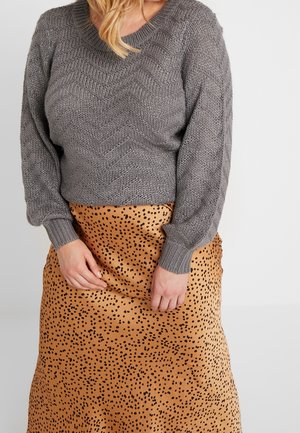 Femme portant un pull en maille texturée grise et une jupe beige avec un motif tacheté noir, debout devant un fond clair uni.