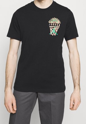 Personne portant un t-shirt Jordan noir avec un petit logo coloré de basket-ball et Jumpman sur la poitrine, associé à un pantalon gris.