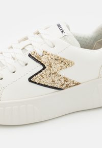 Baskets blanches avec un dessus texturé, présentant un détail zigzag doré pailleté et un fin contour noir. Conception à lacets avec un bout arrondi.